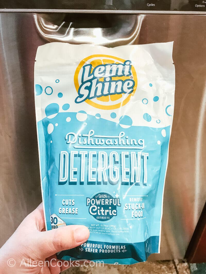 A hand holding up a packet of Lemi Shine Dishwasher Detergent, in front of a dishwasher. 