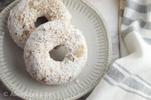 Two powdered apple cider donuts on a plate next to a grey and white towel.