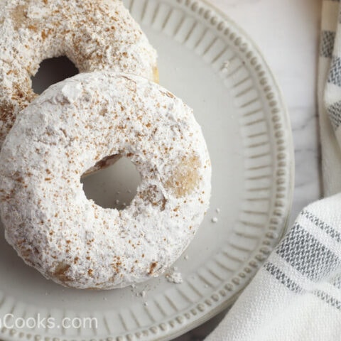 Two powdered apple cider donuts on a plate next to a grey and white towel.