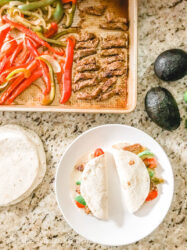 Overhead shot of two fajitas on a white plate above a sheet pan of steak and veggies.