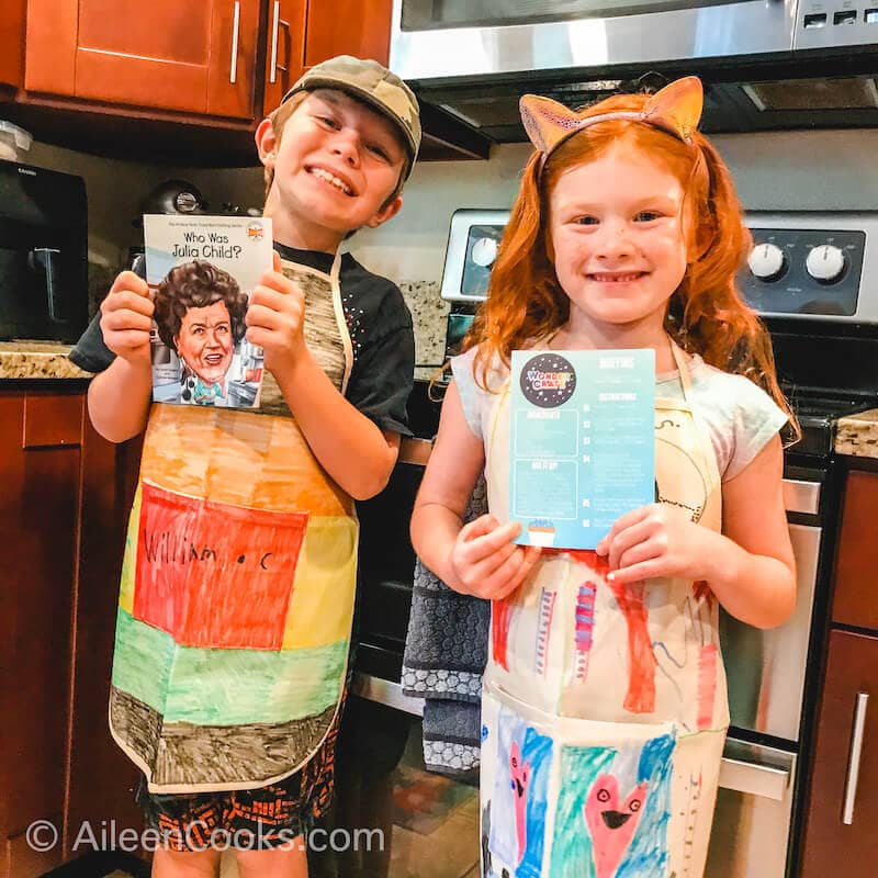 Two kids in colorful aprons smiling and standing in a kitchen.