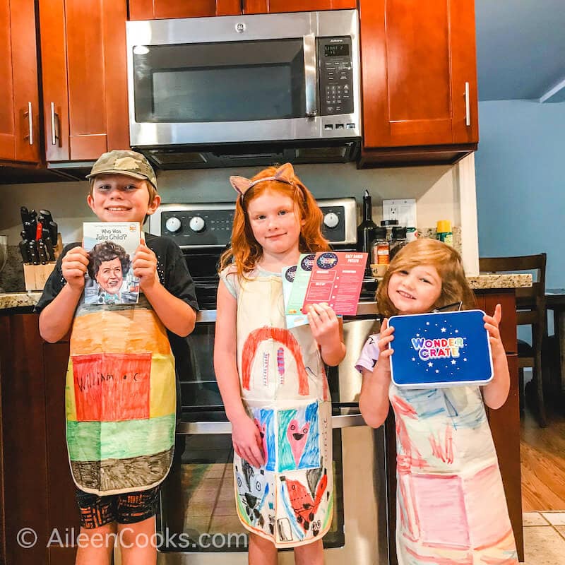 Three kids standing in colorful aprons and standing in a kitchen. 