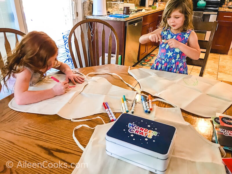 Two girls sitting at a wooden table, decorating aprons with fabric markers.