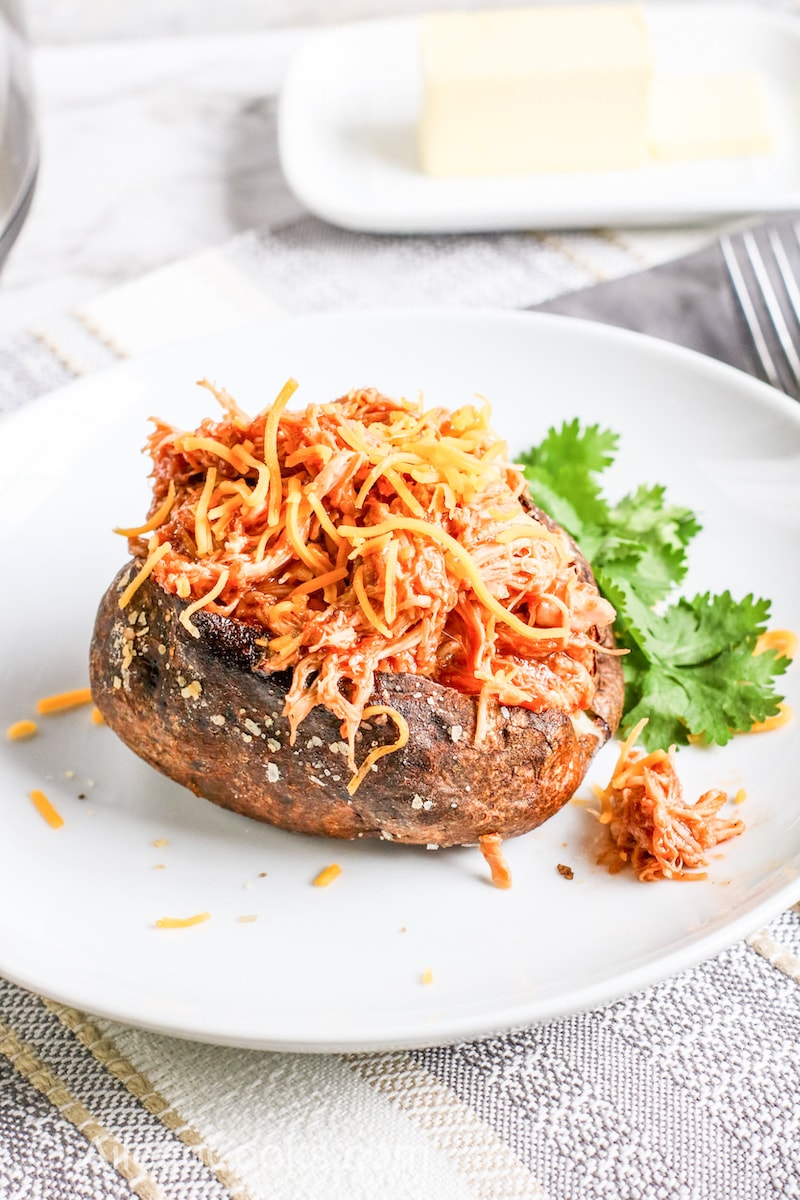 A baked potato on a white plate and topped with shredded chicken and cheese.