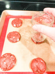 Red Velvet cookies being pressed into crinkle cookies with the bottom of a glass.