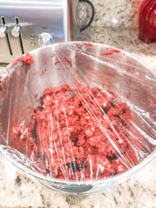 Red velvet cookie dough in a metal mixing bowl and covered in plastic wrap.