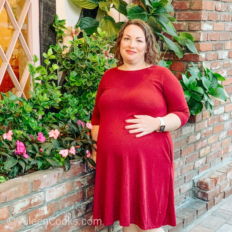 A pregnant woman in a red dress, standing in front of a planter of flowers.