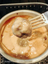 A fork holding a meatball over a cake pan of Swedish meatballs with sauce.