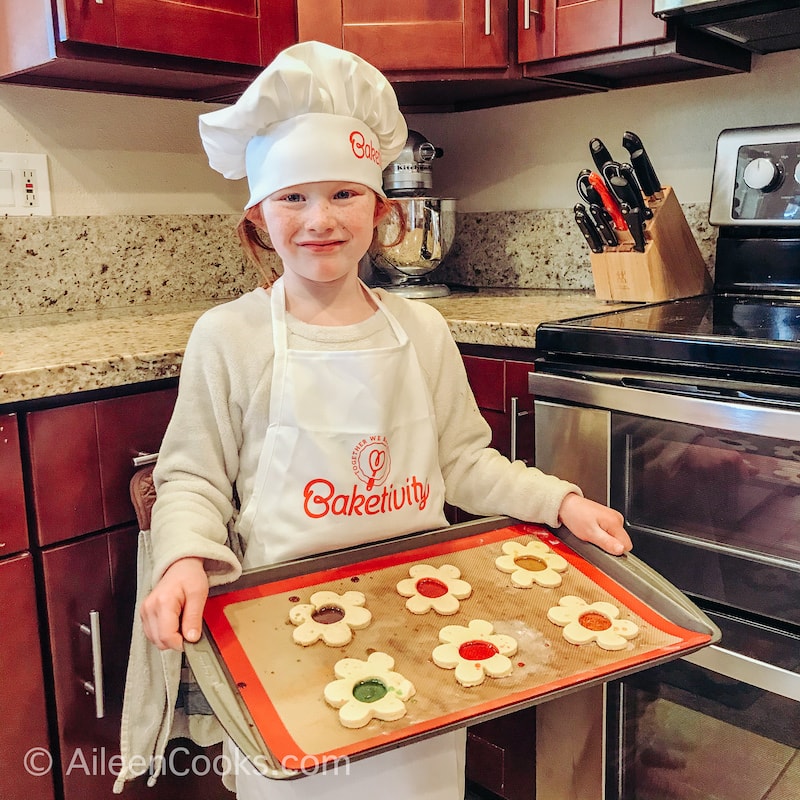 A girl in an apron and chef hat, holding a cookie sheet of baked cookies.
