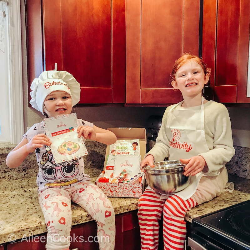 Two girls sitting on a counter, holding a mixing bowl and a recipe card.