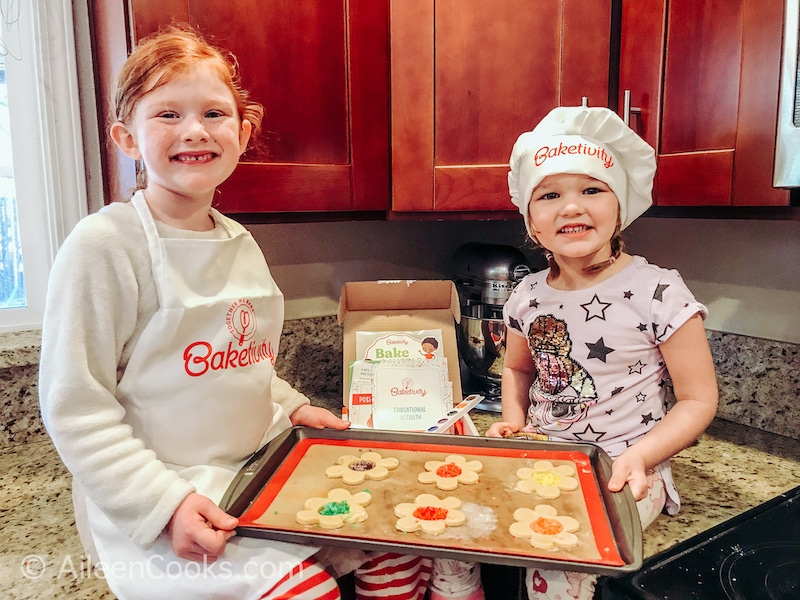 Two girls sitting on a counter holding a cookie sheet of flower-shaped cookies with candy centers.