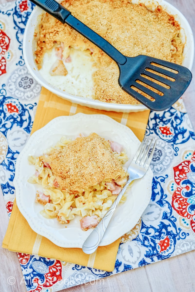 A white casserole dish next to a white plate filled with a large portion of chicken cordon bleu casserole.