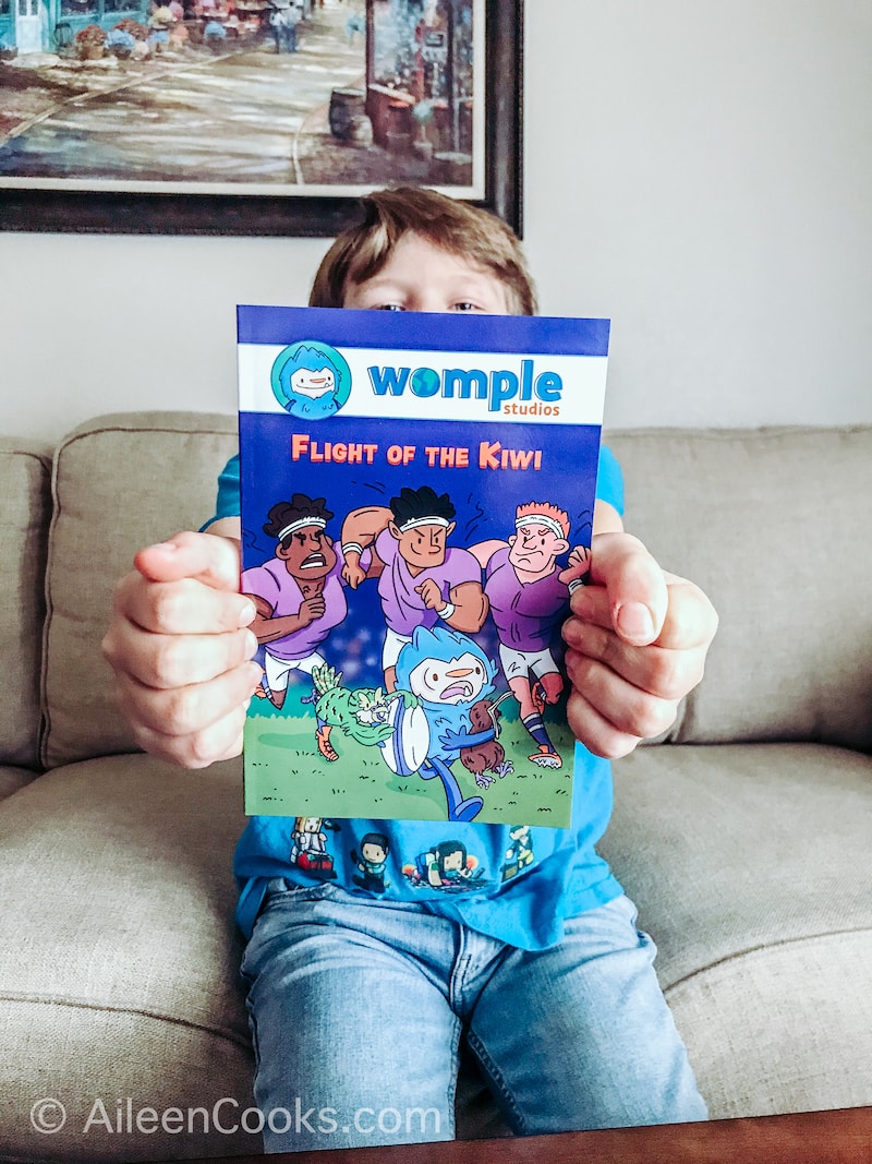 A boy sitting on a beige couch, holding the book Flight of the Kiwi outstretched in front of him.