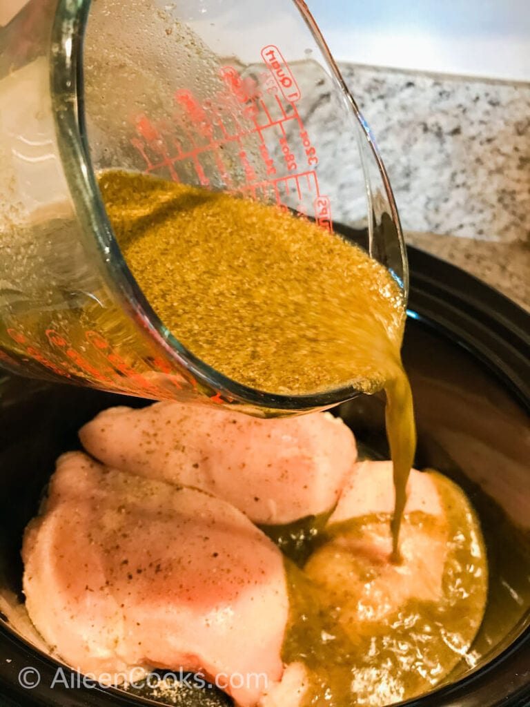 Pesto and chicken broth being poured over chicken inside a slow cooker.