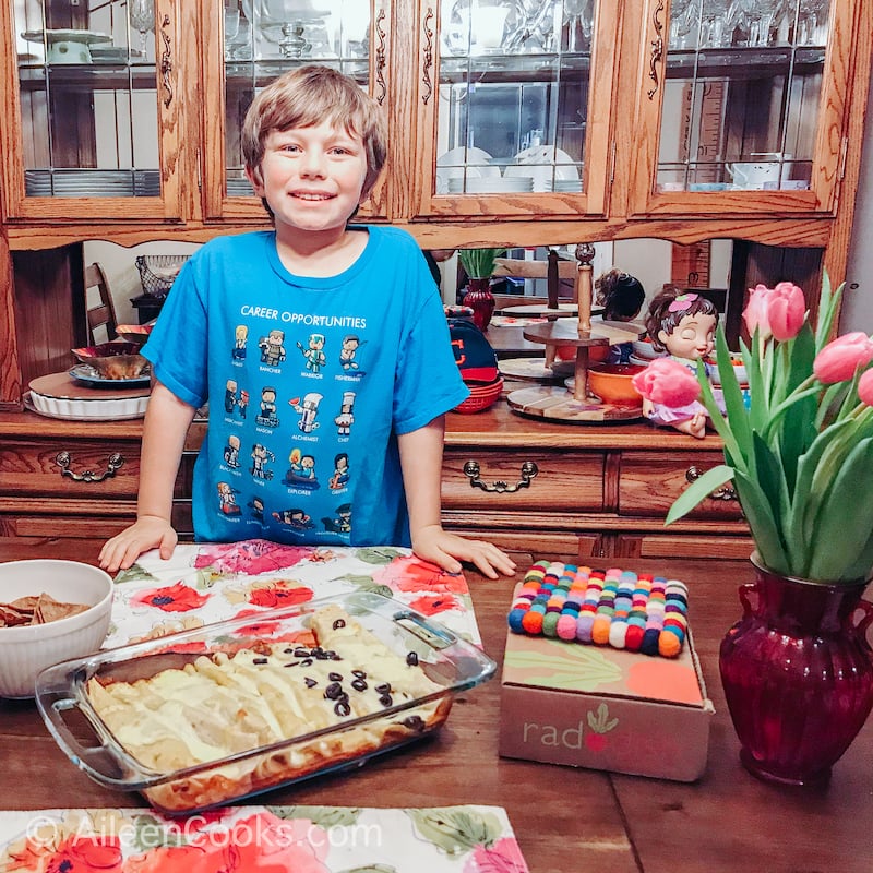 A boy in a blue shirt, standing in front of a dish of enchiladas.