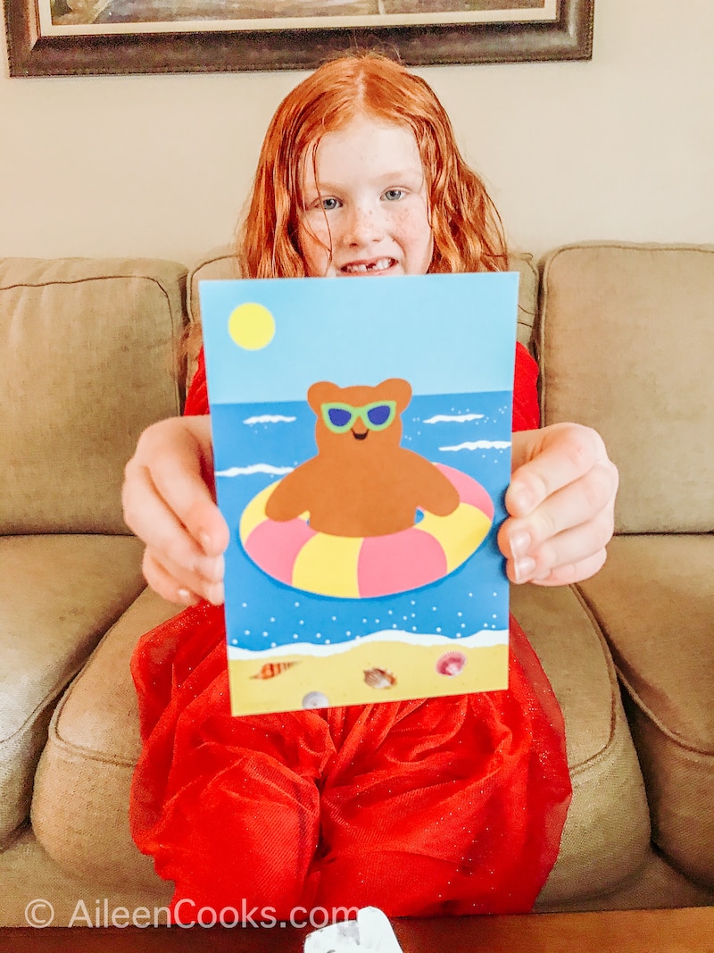 A little girl holding up a postcard with a bear on it.