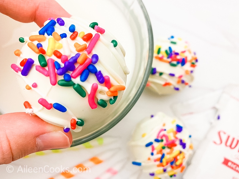 A hand holding a white hot cocoa bomb with rainbow sprinkles.