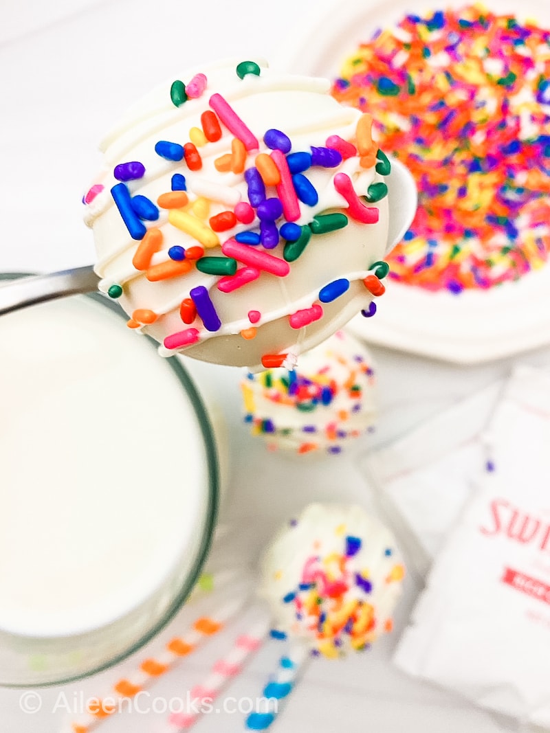 A spoon holding up a white chocolate hot cocoa bombs covered in rainbow sprinkles.