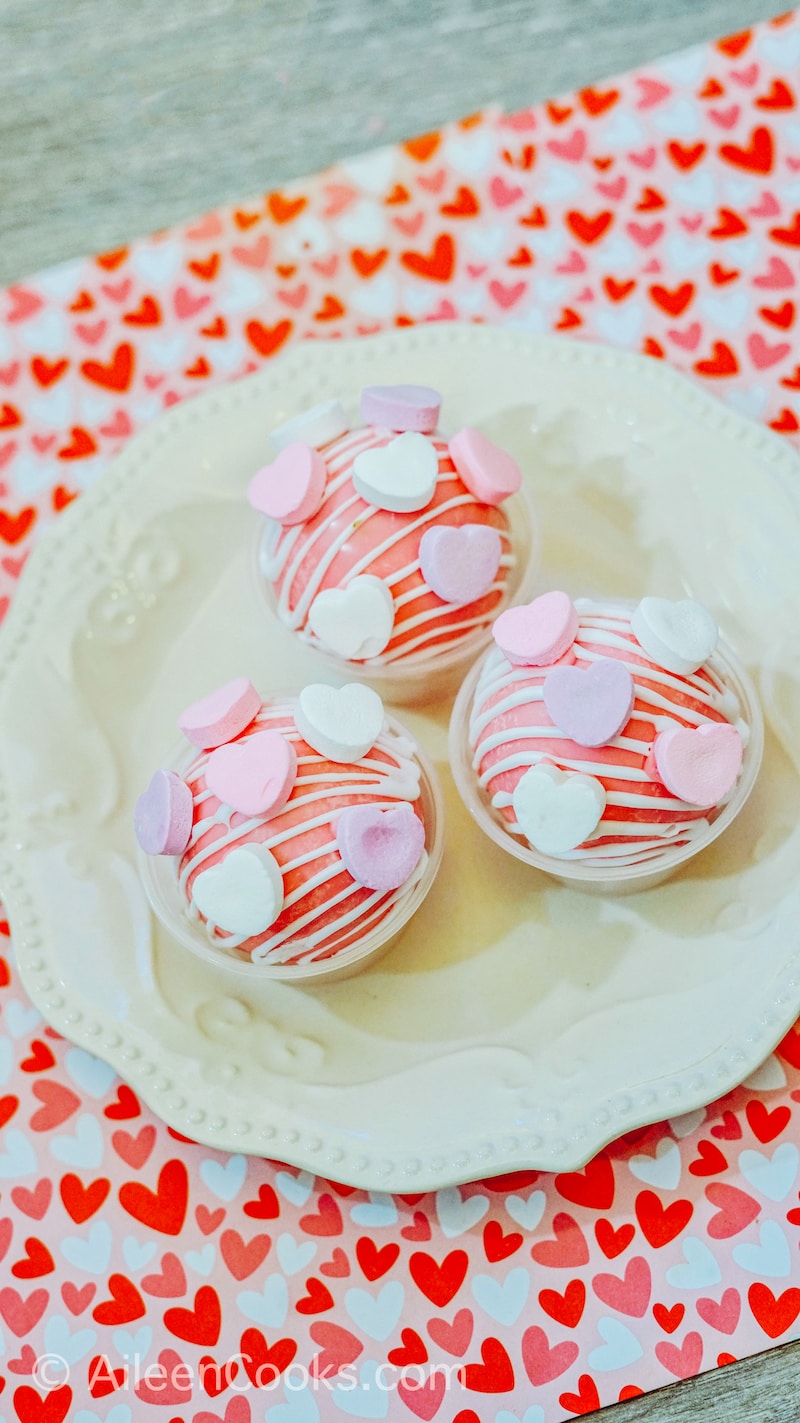A pink and red heart placemat with a white plate of hot cocoa bombs on top.