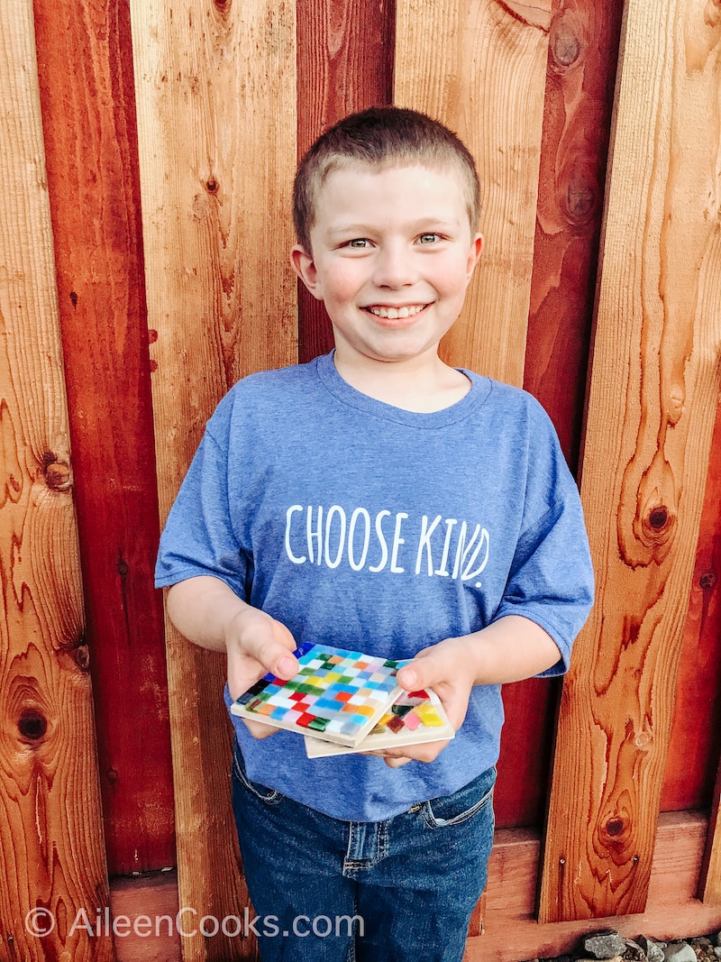 A boy smiling and holding two hand-made mosaic coasters.