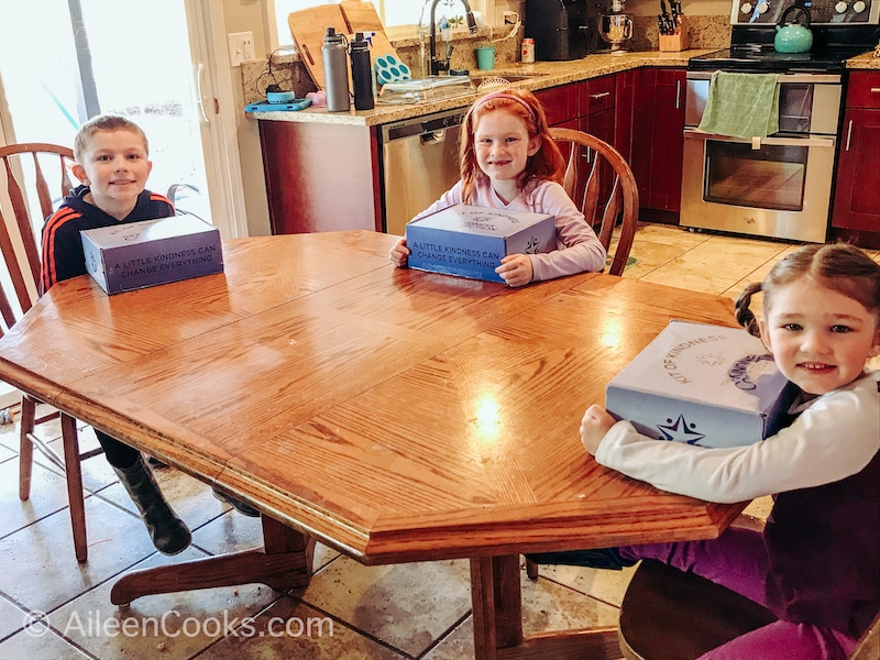 Three kids sitting at a table with kindness kit boxes.