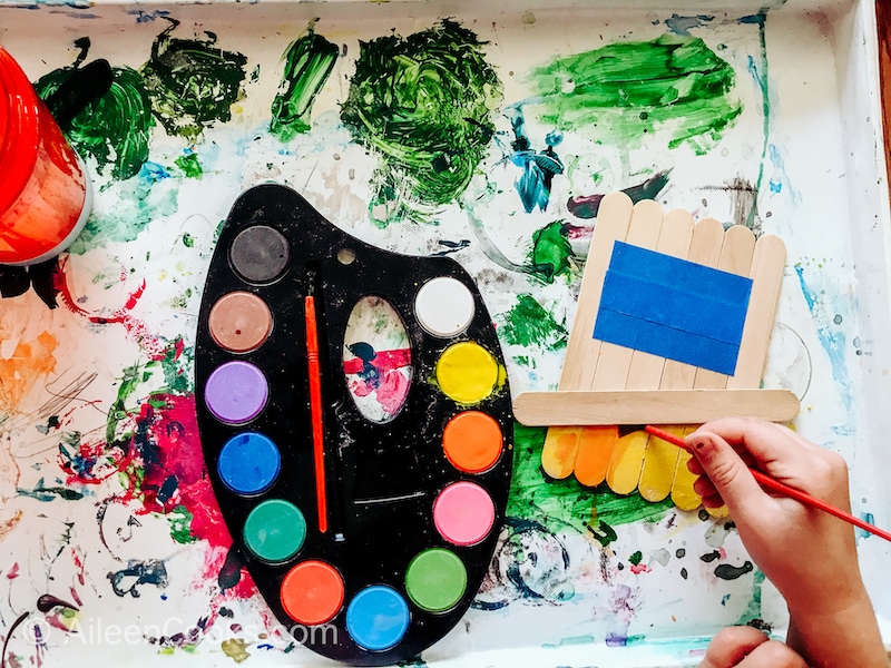 A child's hand painting Popsicle sticks orange.