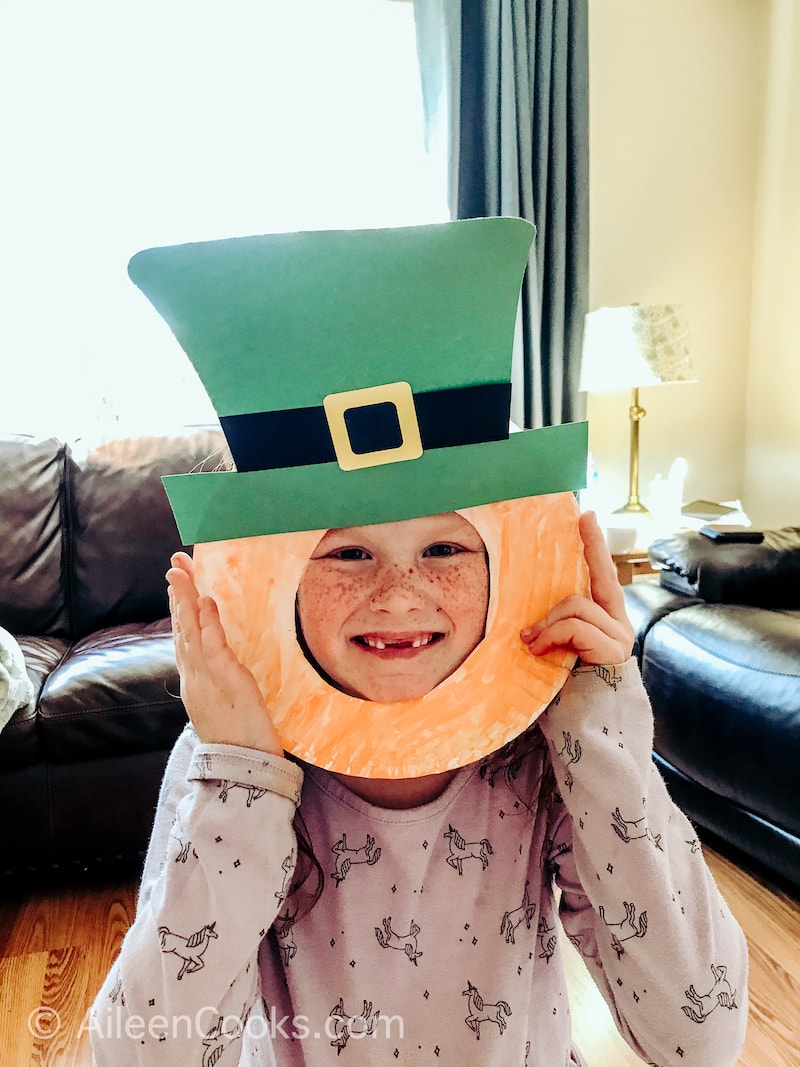 A little girl's face poking through a hand made leprechaun mask craft.