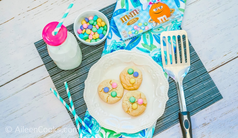 Overhead shot of three Easter cookies on a white plate surrounded by M&Ms, glass bottle of milk, straws, and metal spatula.