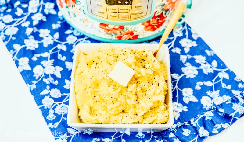 A square bowl of mashed potatoes on top of a blue floral dish cloth.