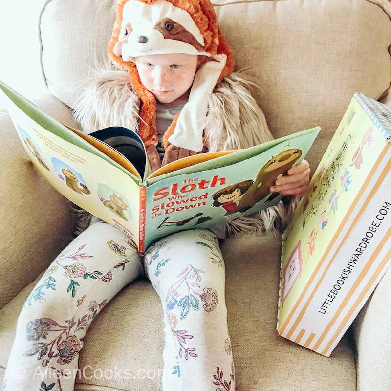 A little girl dressed as a sloth reading a book about a sloth.