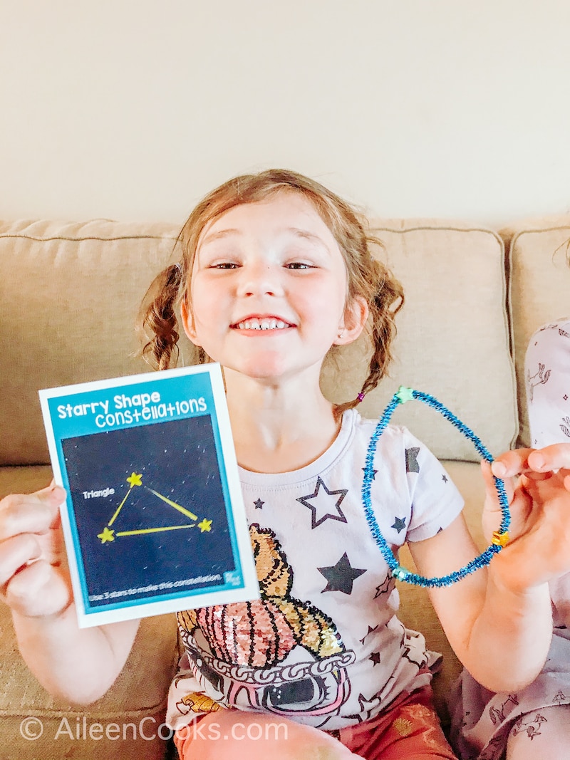 A little girl holding up a picture of a constellation next to her constellation made out of pipe cleaners and star buttons.