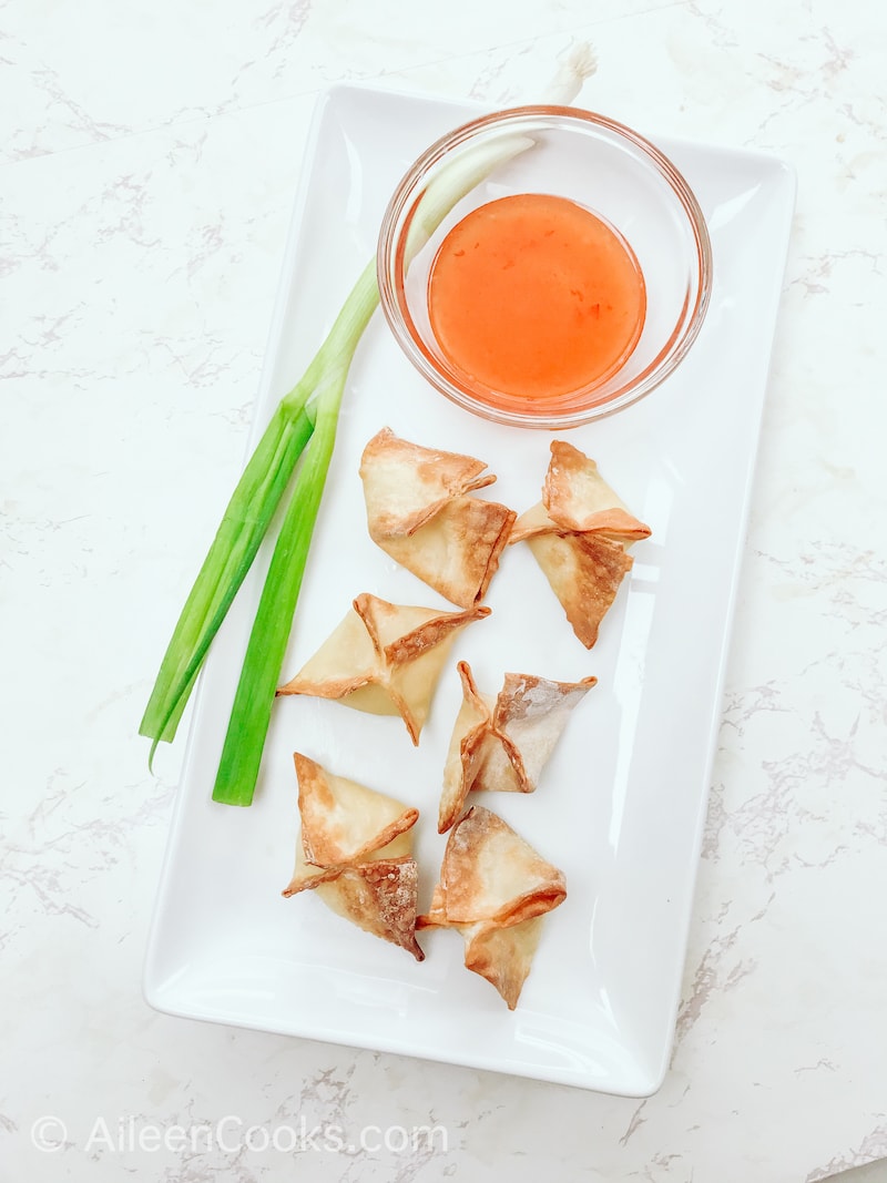 Overhead shot of a white platter with crab Rangoons, small glass bowl of chili sauce, and a green onion. 