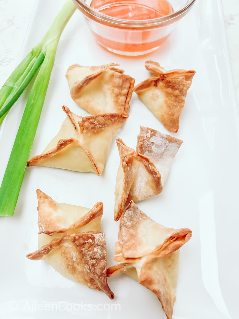 Overhead picture of 6 fried crab rangoons next to a stalk of green onions.