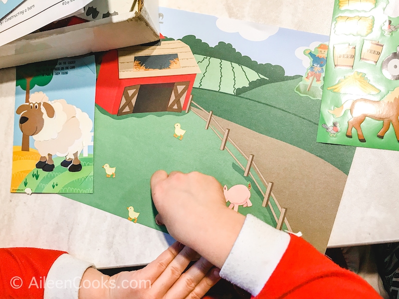 A child's hand putting stickers on a farm scene paper.