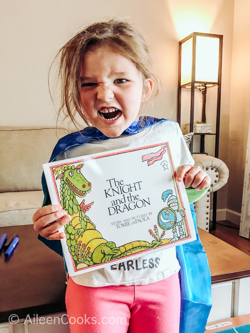 A little girl holding up a book about a dragon and making a scary face.