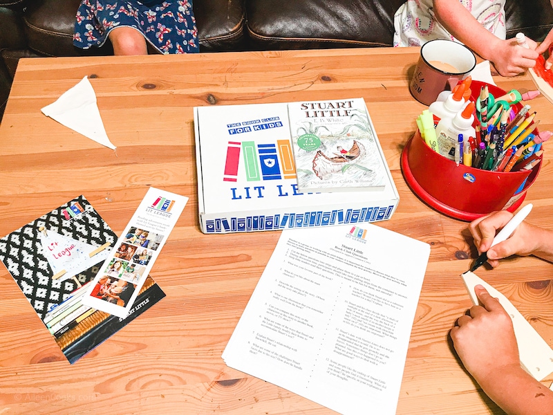 A wood table topped with art supplies, book mark, Lit League Box, discussion questions, and the book Stuart Little.