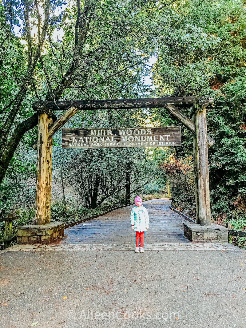 A girl standing under the sign for the Muir Woods National Monument.