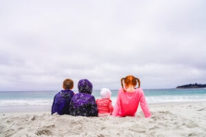 The backs of four kids sitting in the sand at Carmel Beach and looking at the Pacific Ocean.
