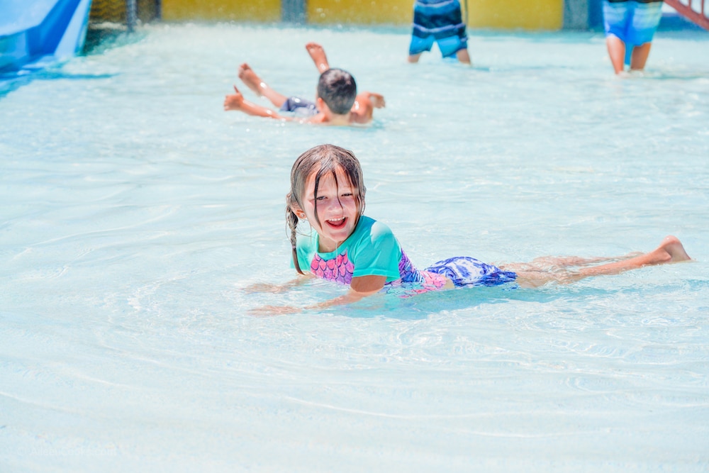 A little girl laying in the shallow water of a water playground and smiling.