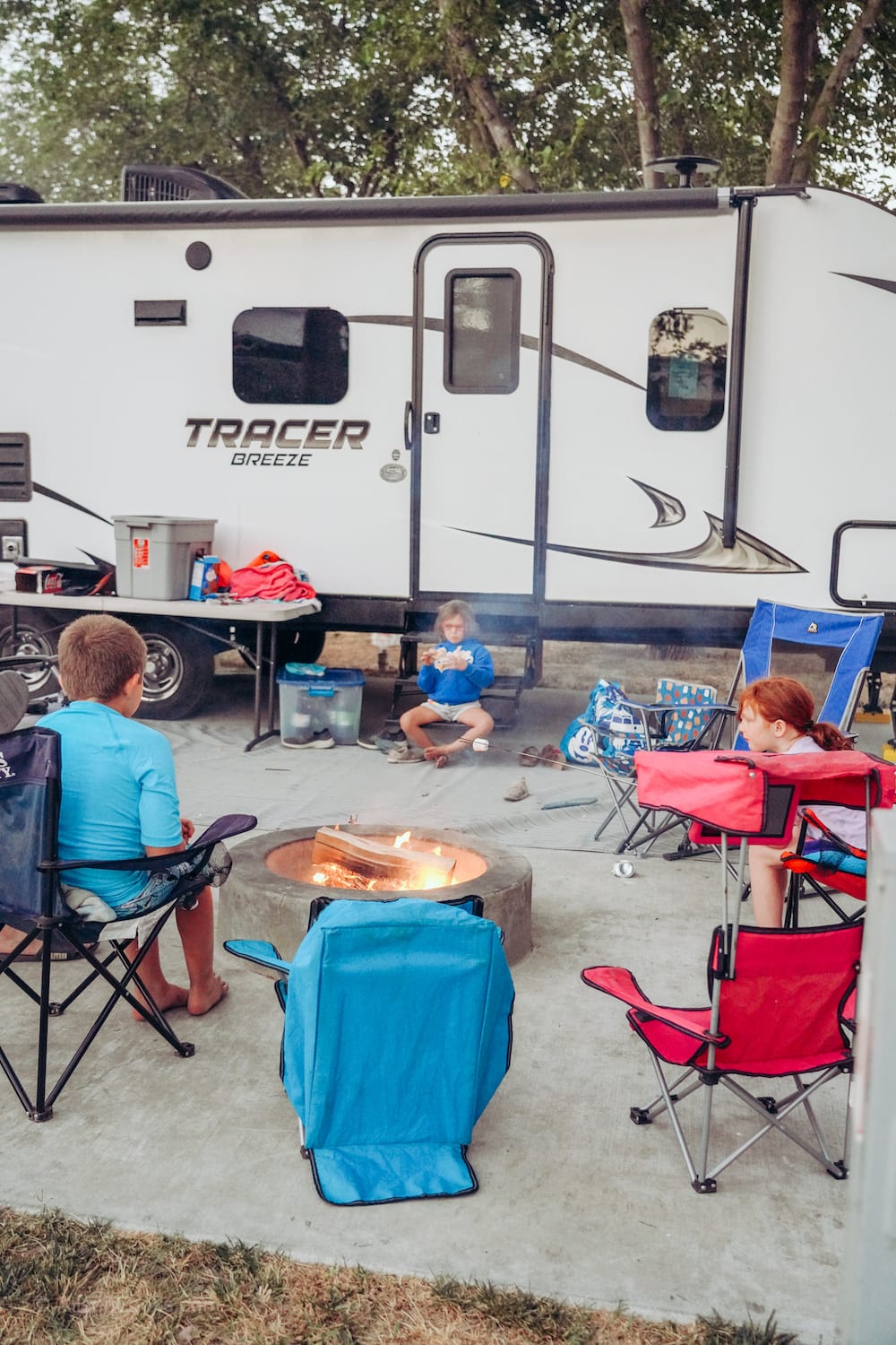 Three kids sitting our a campfire, in front of a travel trailer.