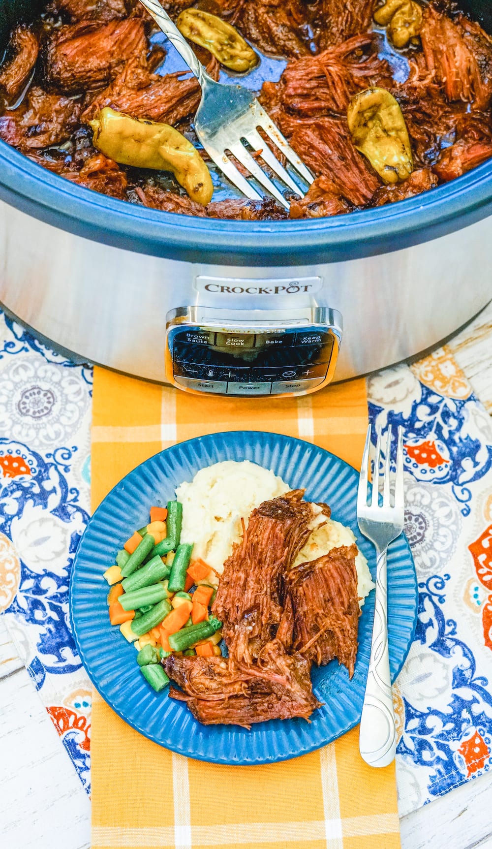 A crockpot filled with Mississippi pot roast behind a blue plate filled with pot roast, mashed potatoes, and mixed vegetables. 