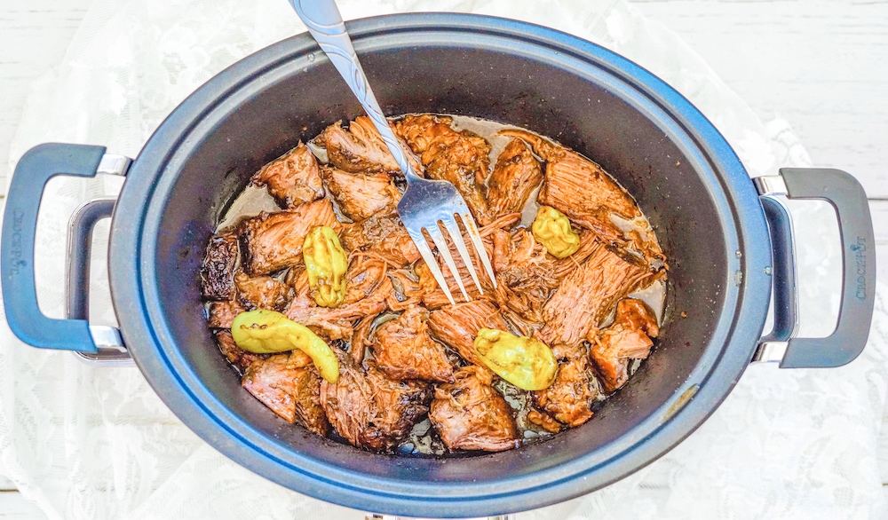 A crock pot filled with cooked pot roast and pepperoncini peppers.