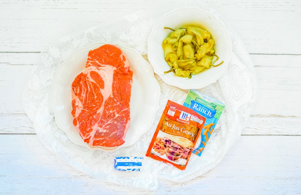 A large platter with a raw chuck roast, seasonings packets, butter, and pepperoncini peppers.
