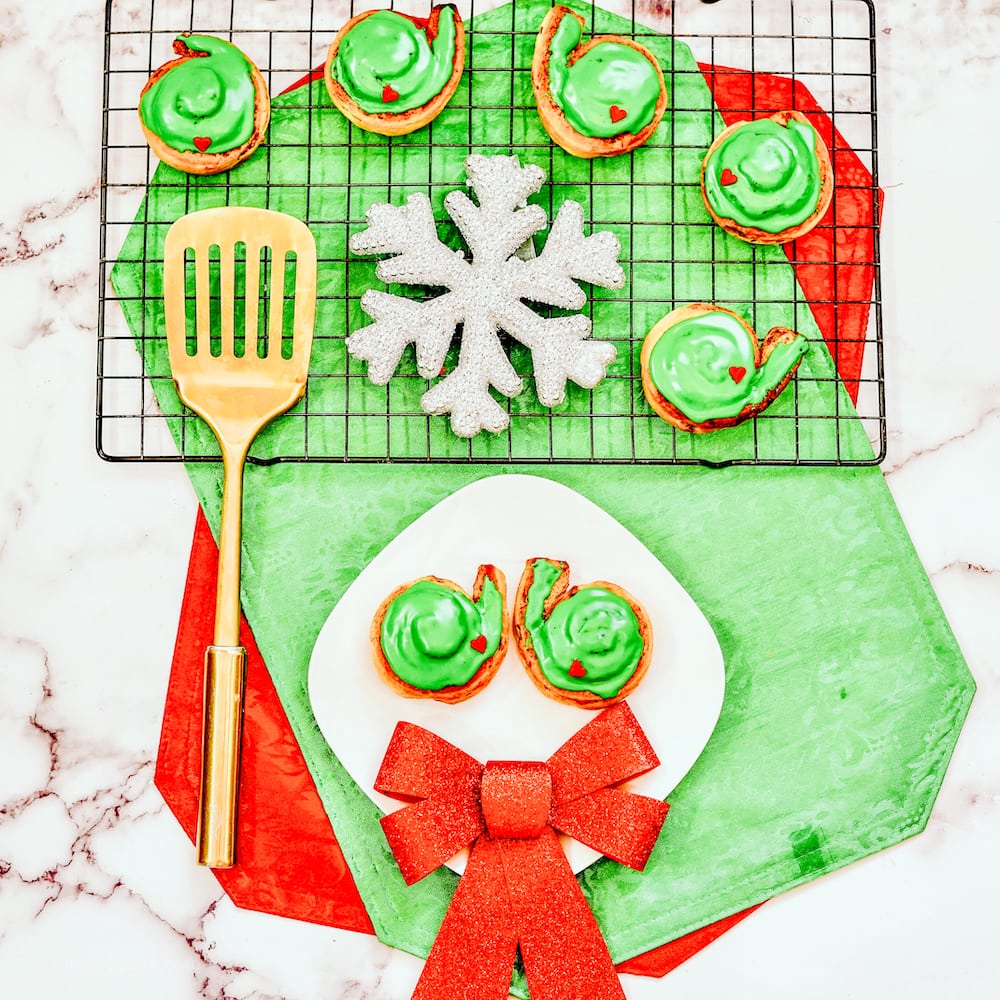A plate of two grinch cinnamon rolls below a cooling rack of green frosted cinnamon rolls with red heart candies.