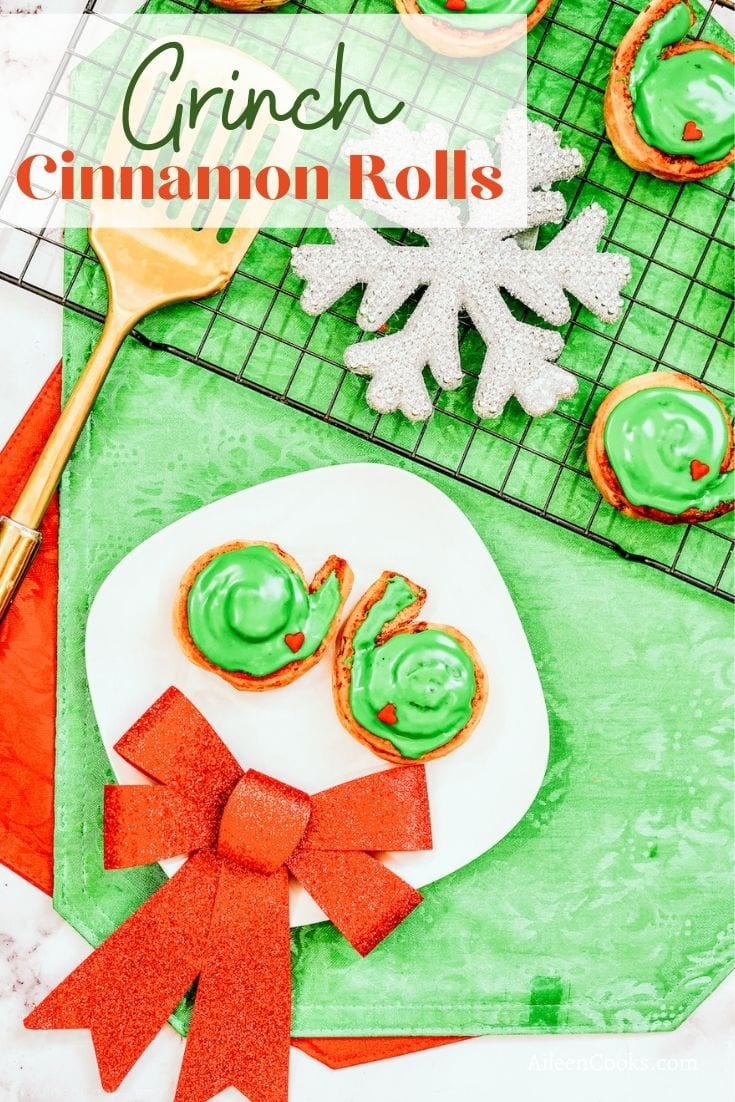 A plate of two Grinch cinnamon rolls over a green placemat with the words "Grinch Cinnamon Rolls" in green and red lettering.