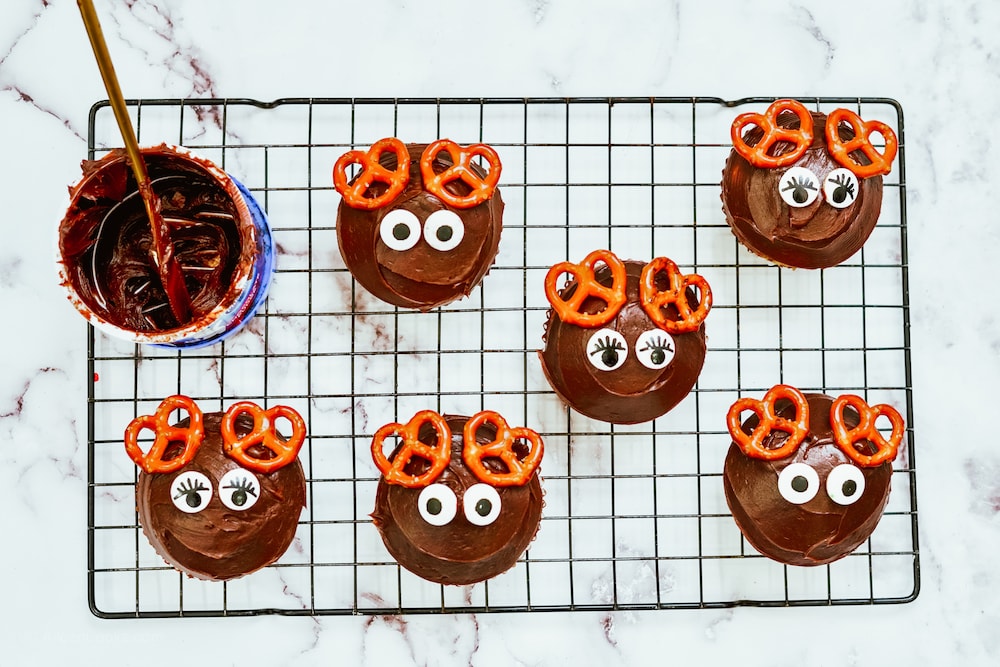 Chocolate cupcakes on a cooling rack with candy eyes and pretzel antlers.