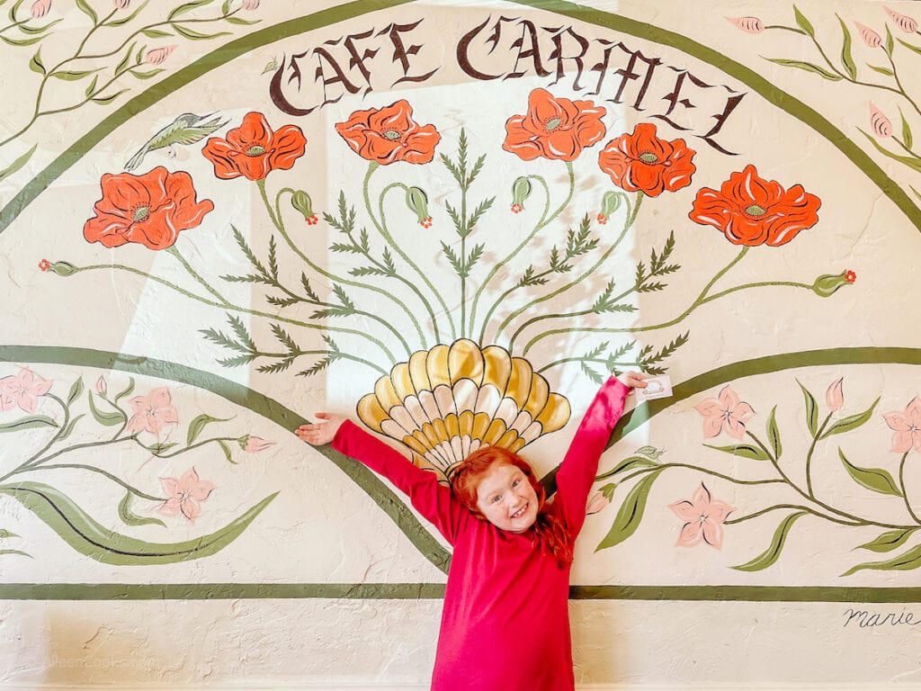 A young girl standing with her arms up in front of a seashell mural that says "Cafe Carmel".