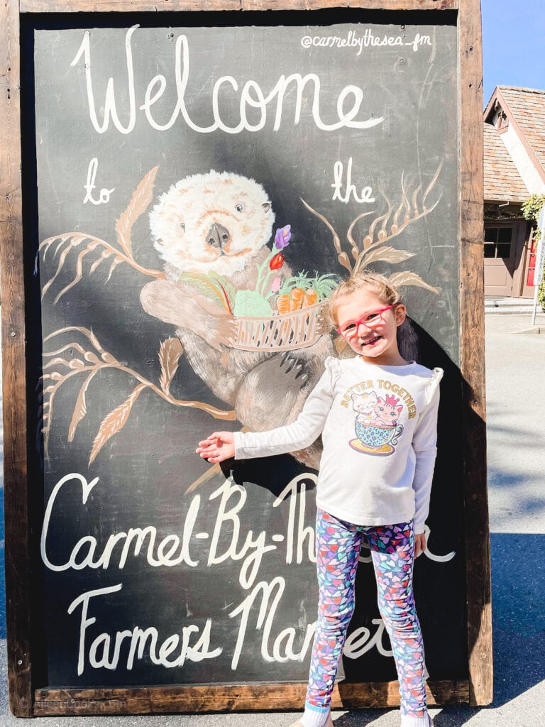 A young girl posing in front of a sign for the Carmel-by-the-Sea Farmer's Market.