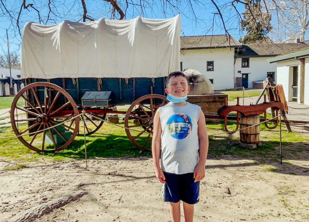 A boy standing and smiling in front of a covered wagon.