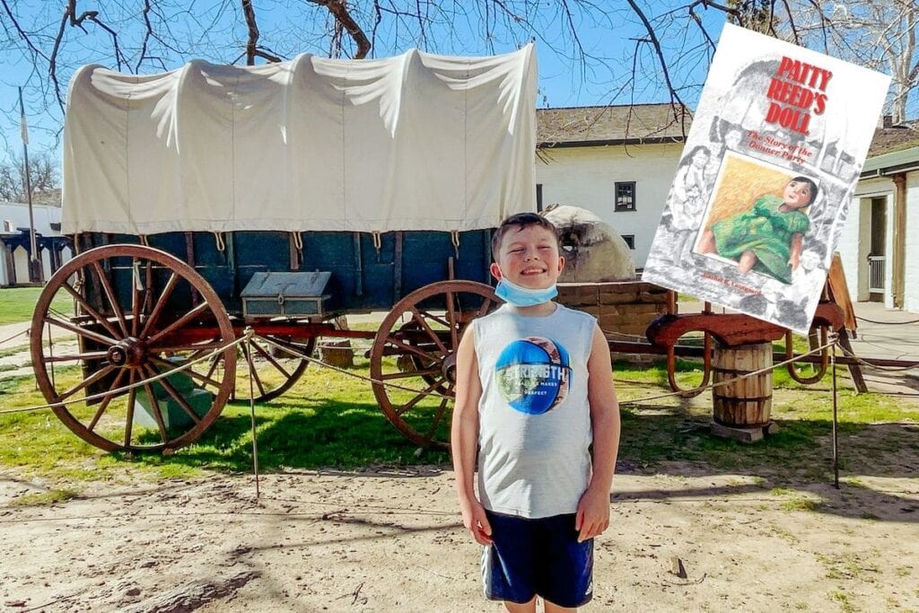 A boy standing in front of a covered wagon at Sutter's Fort with the book cover of Patty Reed's Doll superimposed on top of the image.